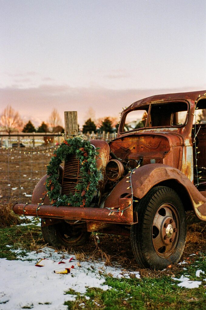 A weathered vintage car adorned with a festive wreath stands in a snowy field at sunset.