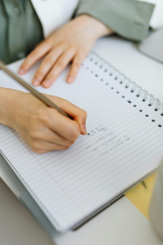 Close-up photo of a person writing in a spiral notebook with a pencil on white lined paper.
